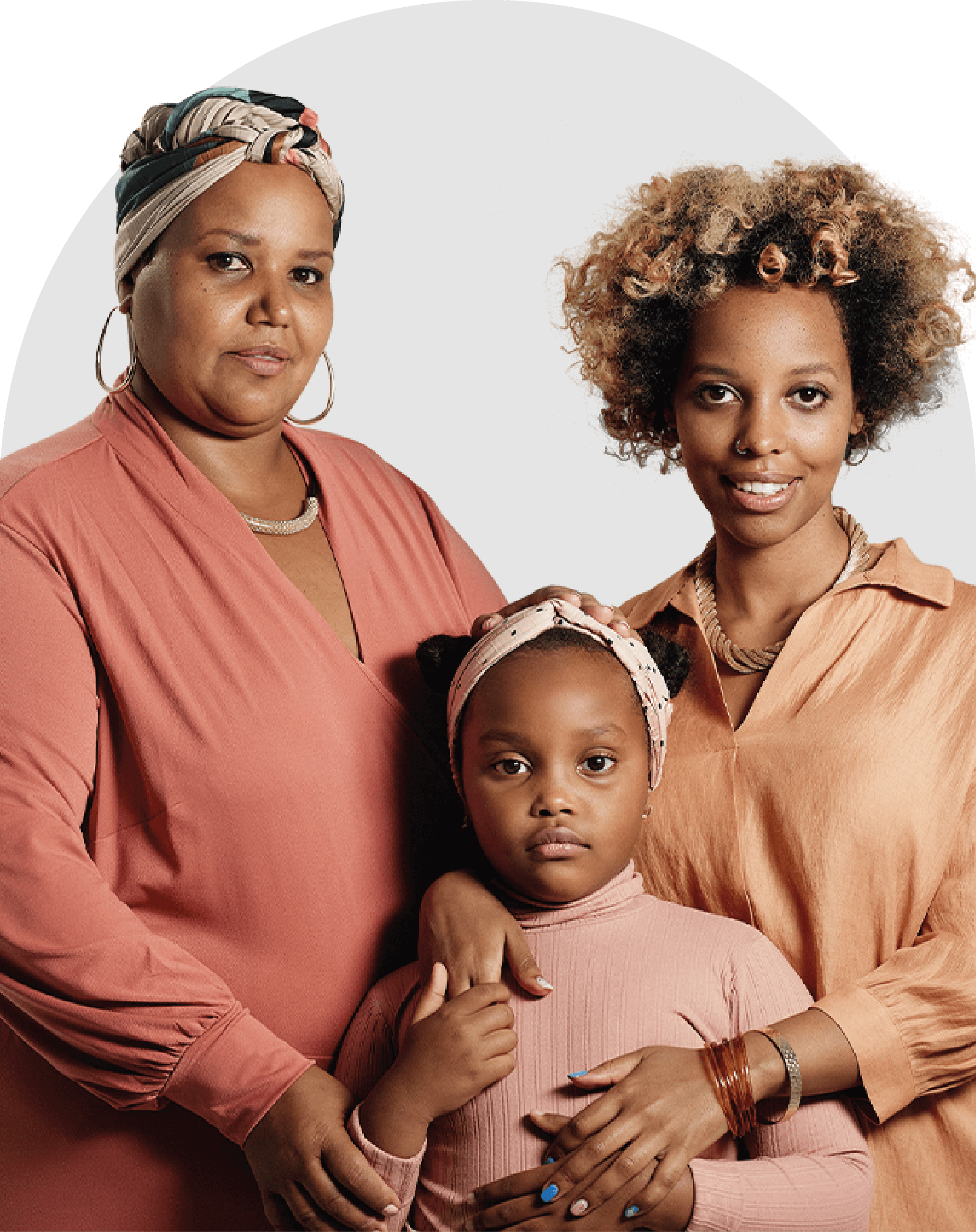 Foto de três mulheres negras, de três gerações diferentes. A primeira, mais velha, usa um turbante na cabeça com um vestido coral, a segunda usa os cabelos soltos com uma camisa laranja e a terceira é uma criança que usa roupas rosas.