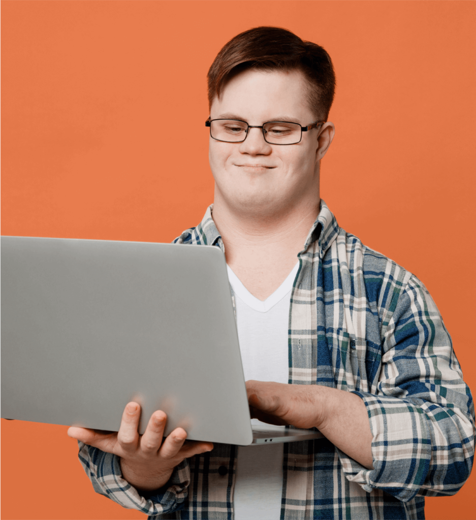 Foto de um homem branco com síndrome de down. Ele usa óculos, uma camiseta branca sobreposta com uma camisa xadrez e segura um computador.
