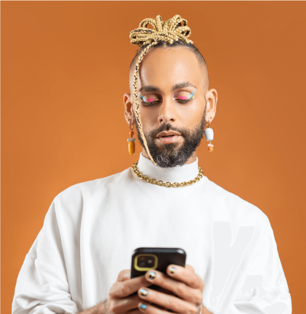 Foto de um homem negro de pele clara. Ele usa tranças loiras amarradas em um coque, barba, maquiagem colorida, brincos e uma blusa branca de gola alta. Ele está segurando um celular.